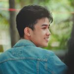 A young man with a joyful expression wearing a denim jacket in a natural setting.